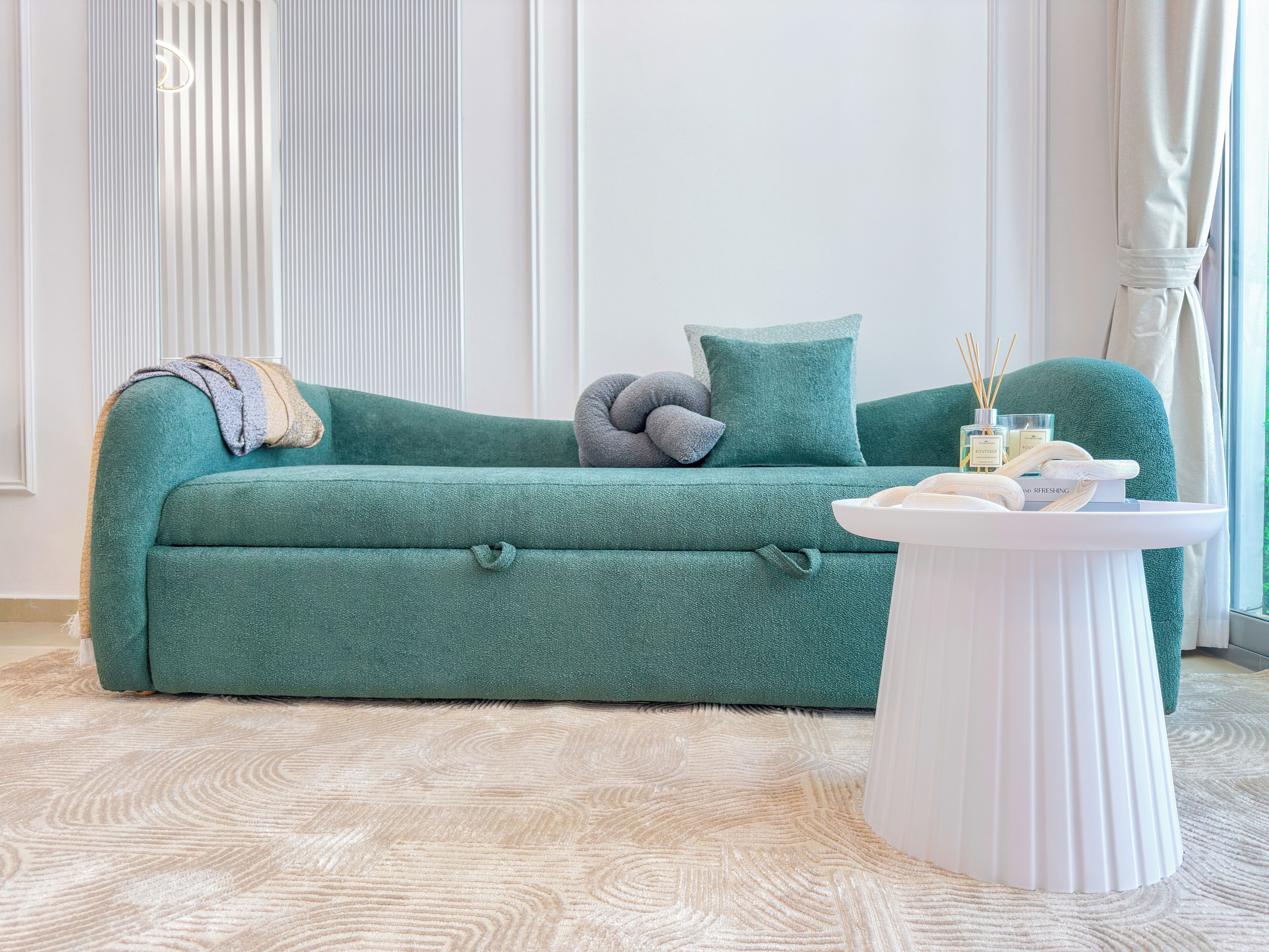 Een teal kleurige bank met een witte bijzettafel, een grijze kussen en een witte vaas met kaarsen.
