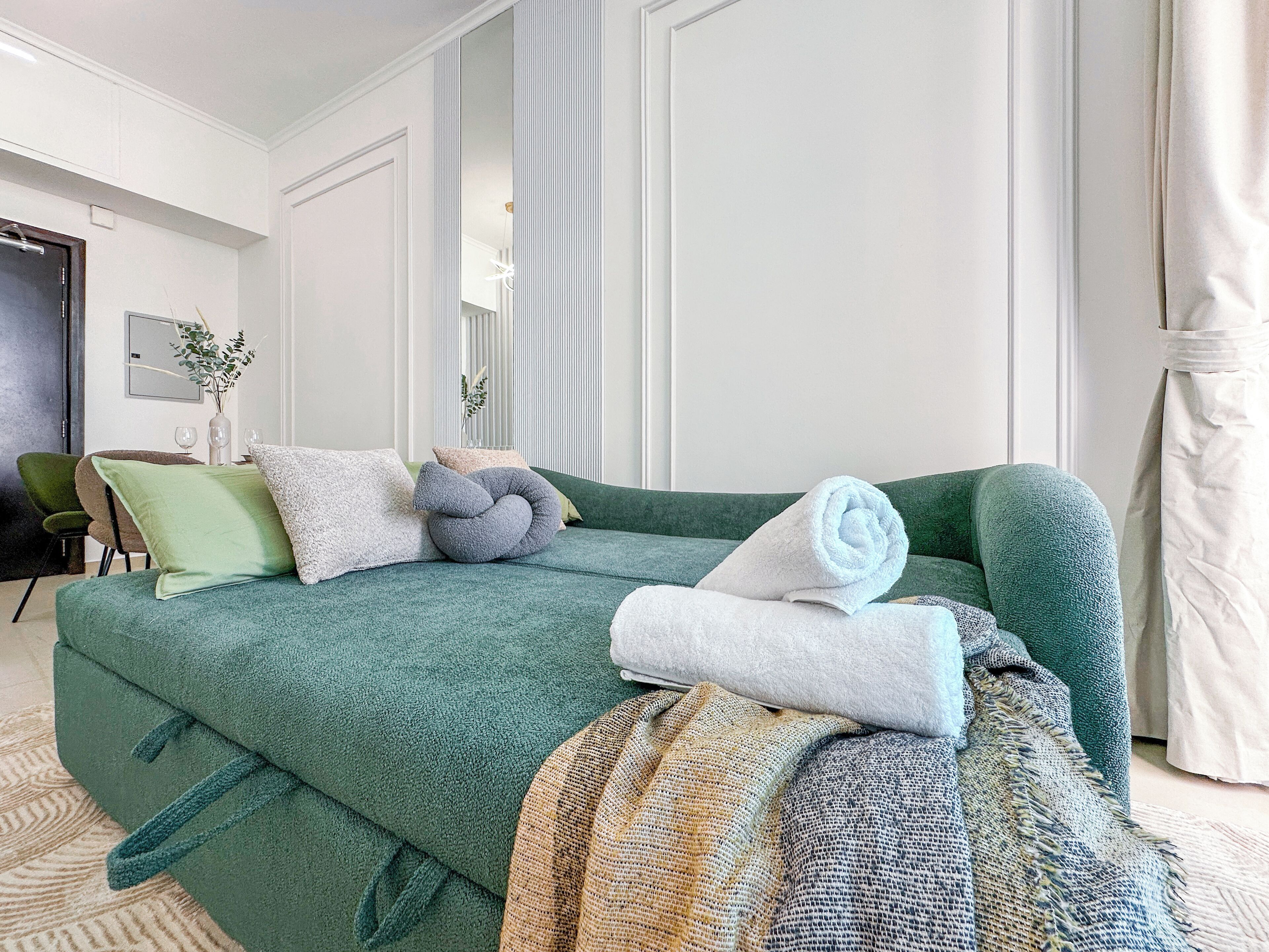 A green sofa with rolled white towels, a patterned throw, and multiple pillows in a well-lit room with a door, a chair, and a vase with plants.