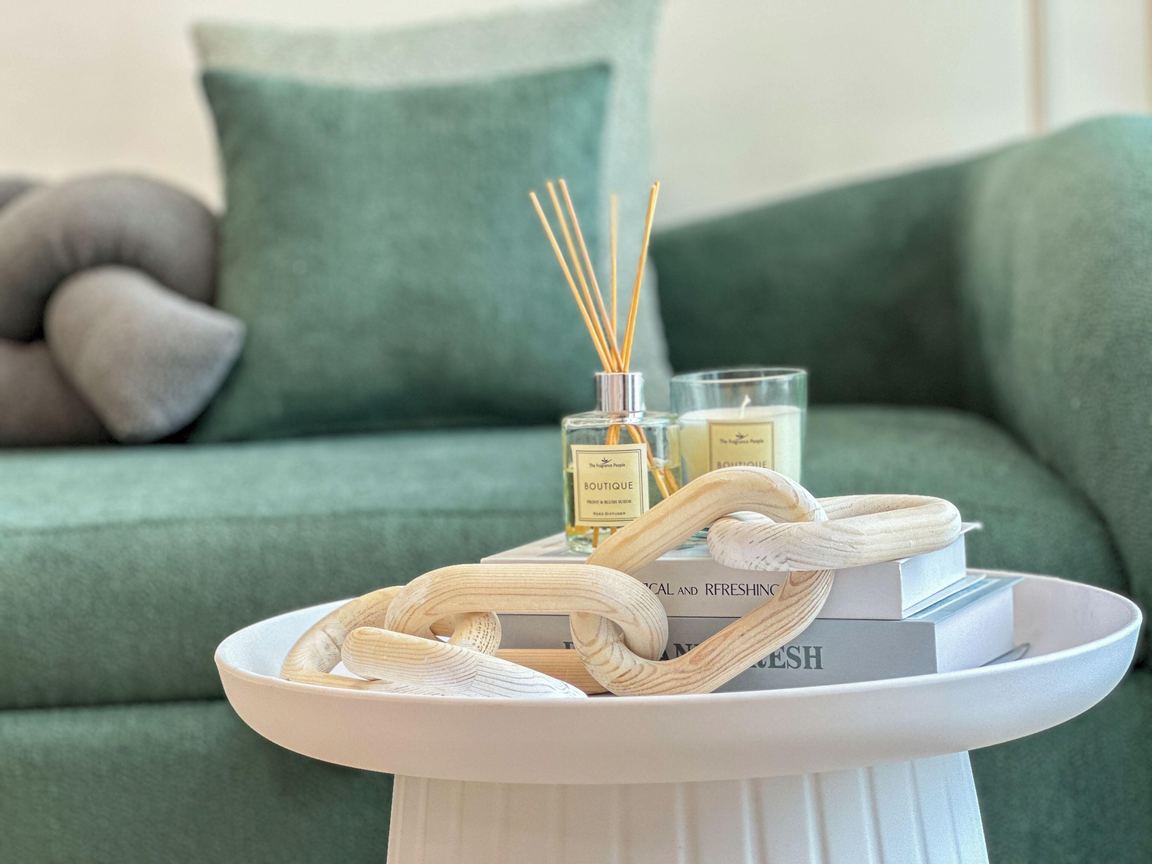 A white tray with a woven basket, two bottles of perfume, and a candle on a white table in front of a green sofa with a pillow.