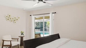 A bedroom with a bed, a chair, a small table with a plant, a ceiling fan, and a view of a pool through a sliding door. - Heated Pool/ 4 Bedrooms (Naples)