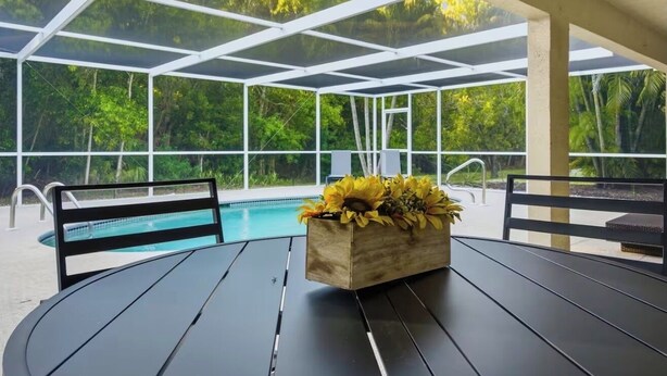 A covered outdoor area with a pool, wooden chairs, and a table with a flower arrangement. - Heated Pool/ 4 Bedrooms (Naples)