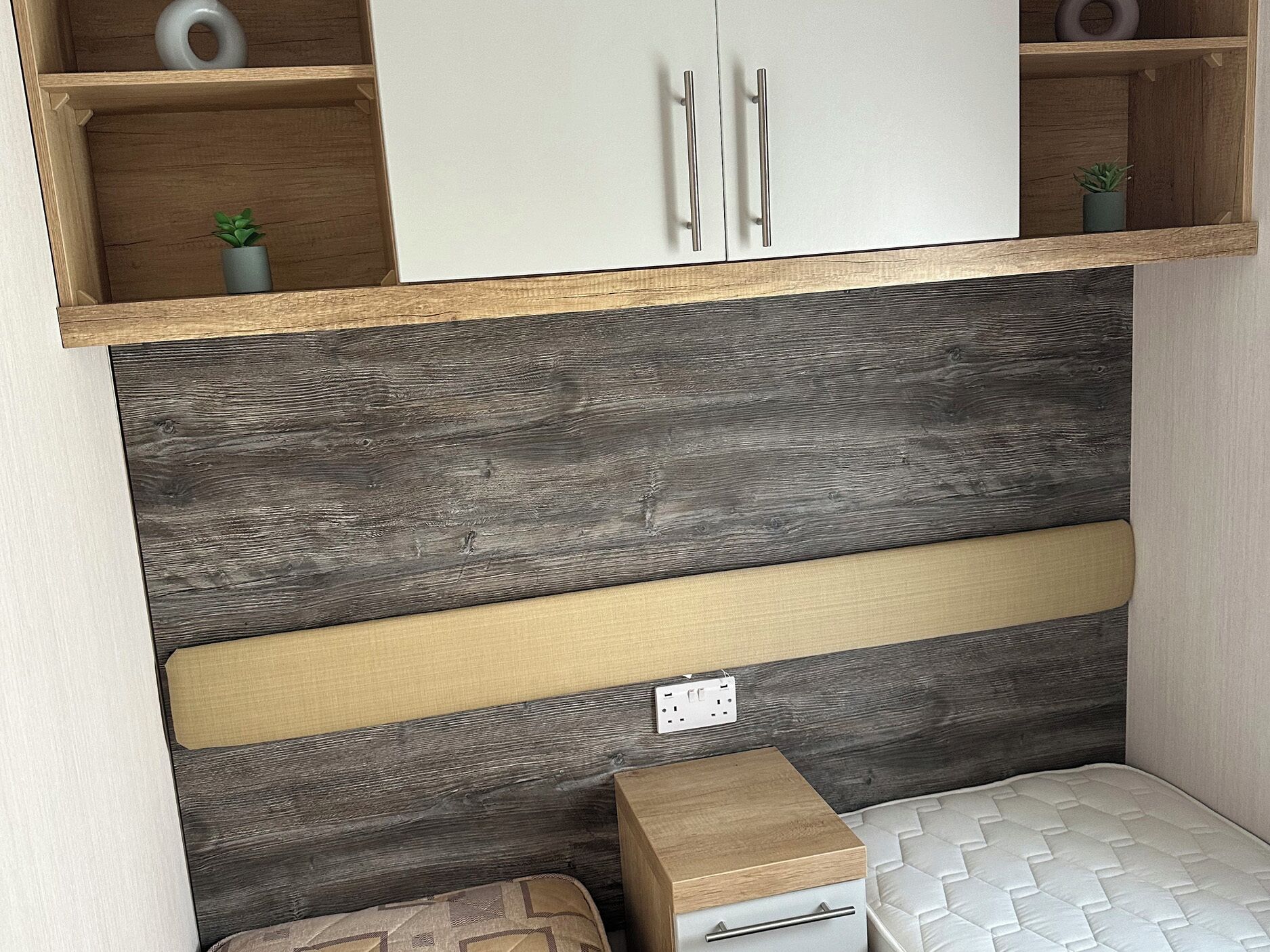 Une chambre à coucher de taille réduite, avec une tête de lit en bois, une petite table de chevet, un matelas et un placard au-dessus.