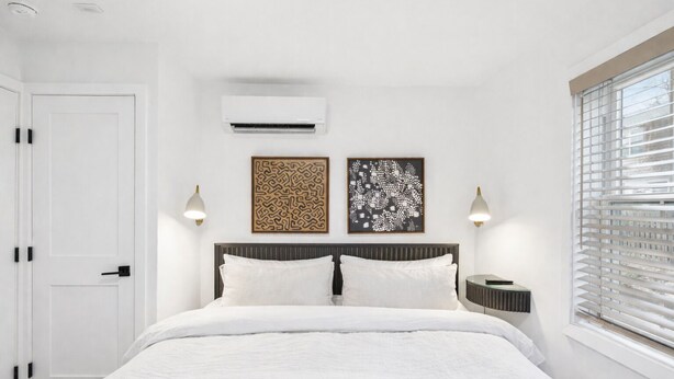 A bedroom with a bed, two wall-mounted air conditioners, a nightstand, a bedside table, and a window with blinds.