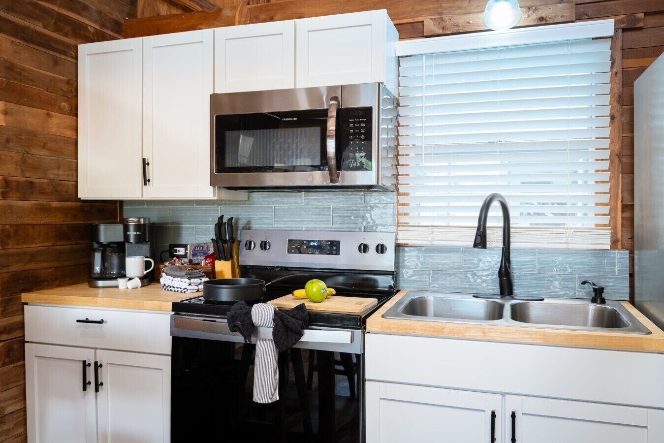 A compact kitchen with white cabinets, a microwave, a stove, a sink, and a window with blinds.