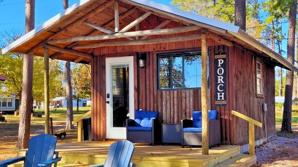 A rustic wooden cabin with a porch, two blue chairs, and a small table. - Mid Lake Campground (Hemphill)
