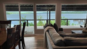 A living room with a large window overlooking a body of water, a wooden dining table with chairs, and a sofa. - Mid Lake Campground (Hemphill)