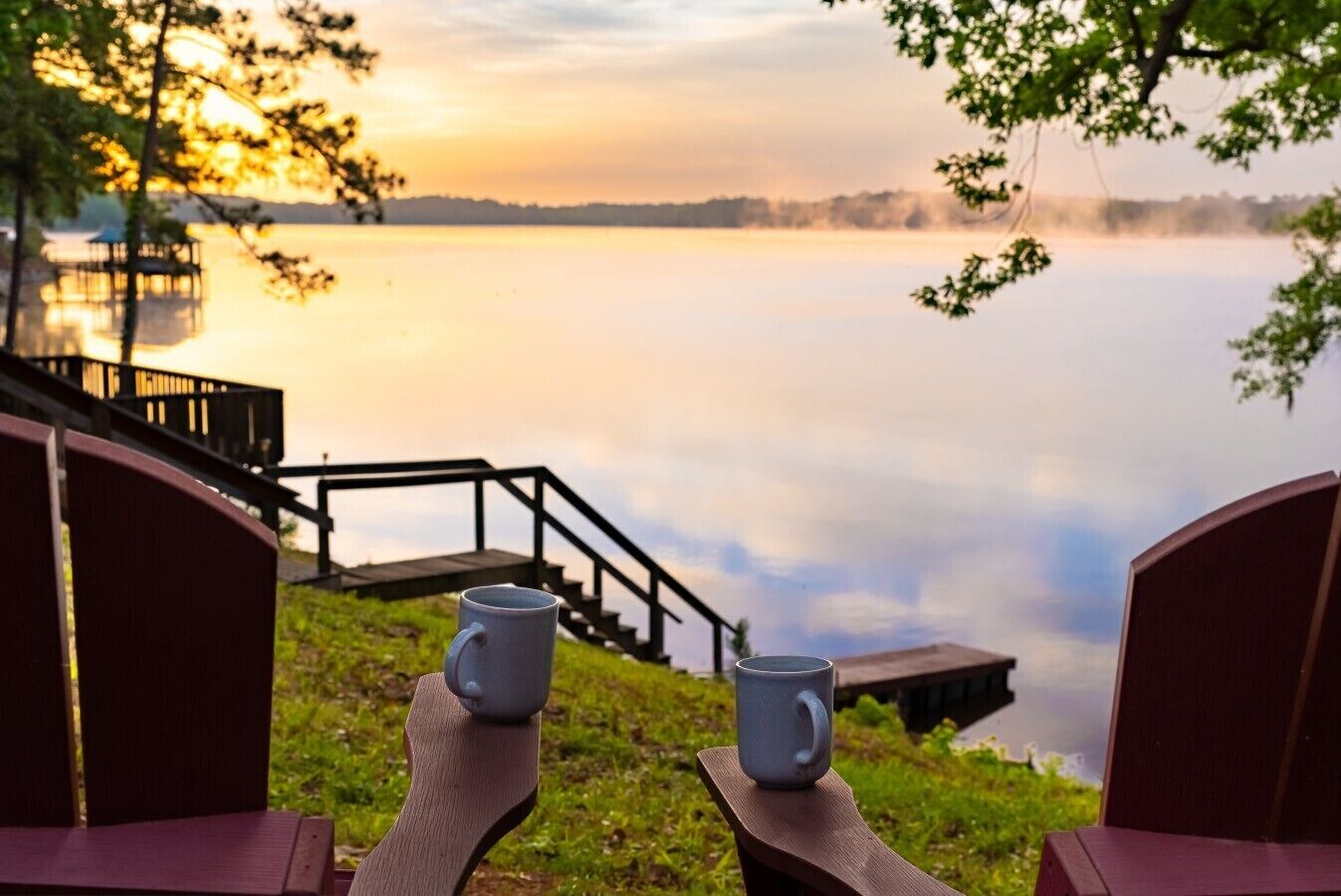 A lakeside view with a wooden deck, two chairs, and two mugs.