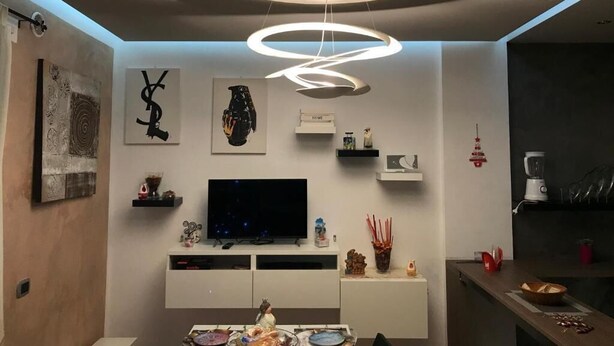 A modern living room with a flat-screen TV, a dining table with a decorative centerpiece, and a unique circular ceiling light fixture. - Elena's house, is a dream retreat for every type of guest (Sannicandro di Bari)