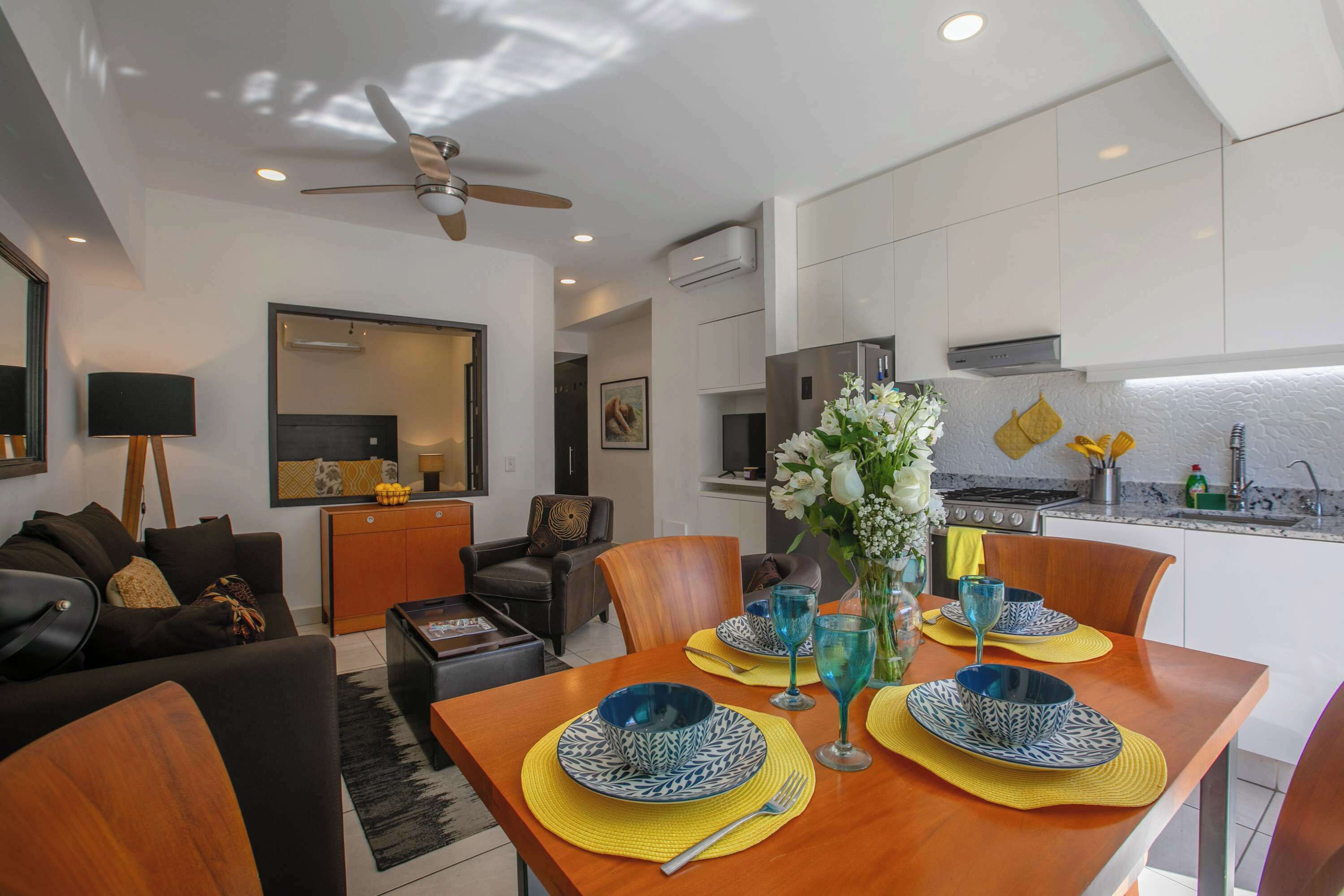 A modern living room with a dining area, featuring a wooden dining table set with blue and white plates, a ceiling fan, and a kitchen with white cabinets and a stove.
