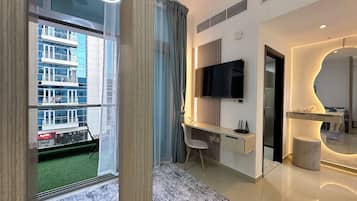 Een moderne woonkamer met een groot raam, een flatscreen televisie aan de muur, een bureau met stoel, en een decoratieve spiegel.