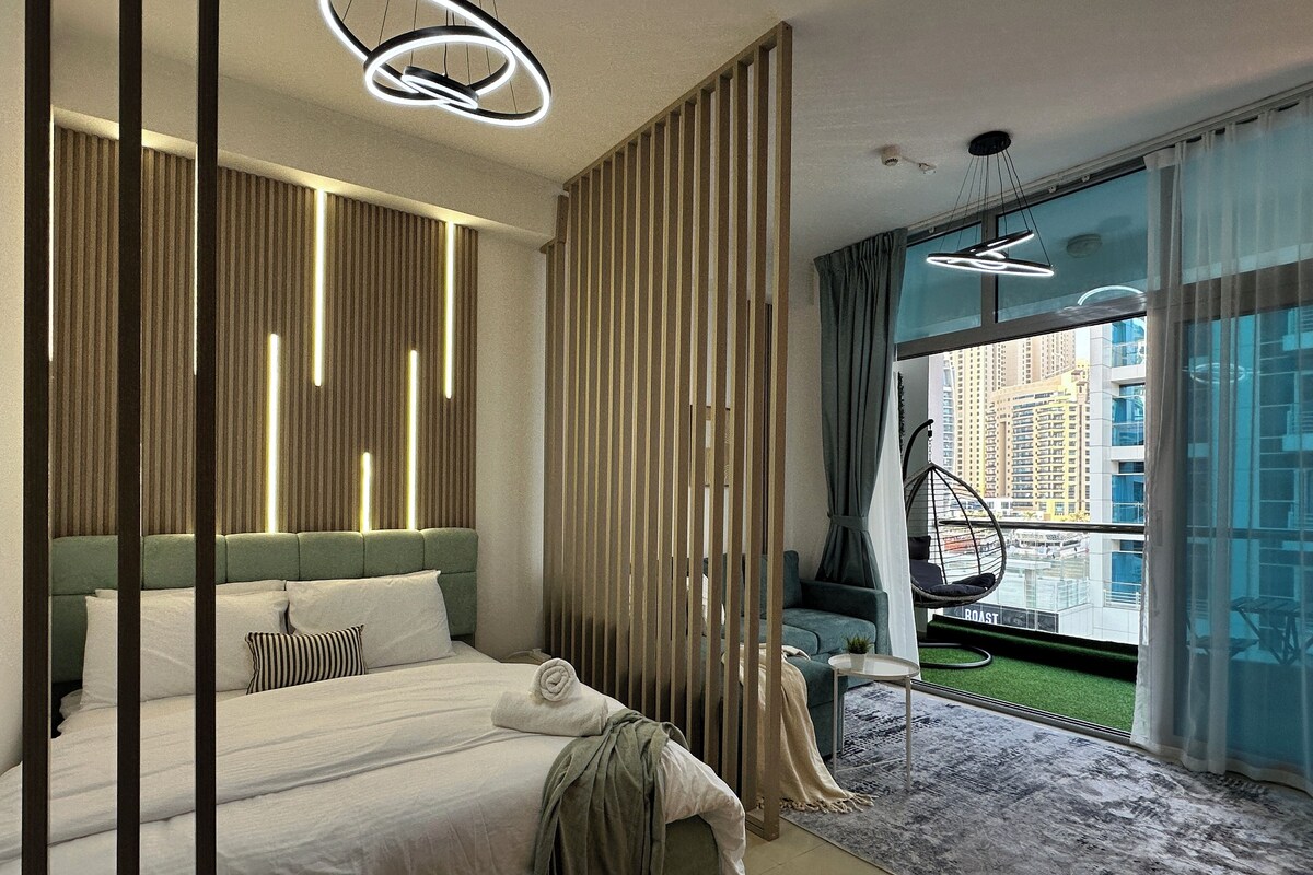 A modern hotel room with a large bed, a hanging chair, a view of the city, and a balcony with a green lawn.