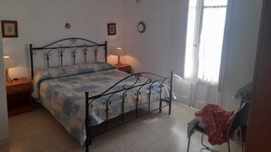 Ein Schlafzimmer mit einem Metallbettgestell, einem Nachttisch mit Lampe, einem Sessel und einem Fenster mit Vorhängen.