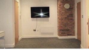 A room with a flat-screen TV mounted on the wall, a brick accent wall, and a carpeted floor. - Cosy 3-Bedroom Family Apartment - Free Parking (Birmingham)