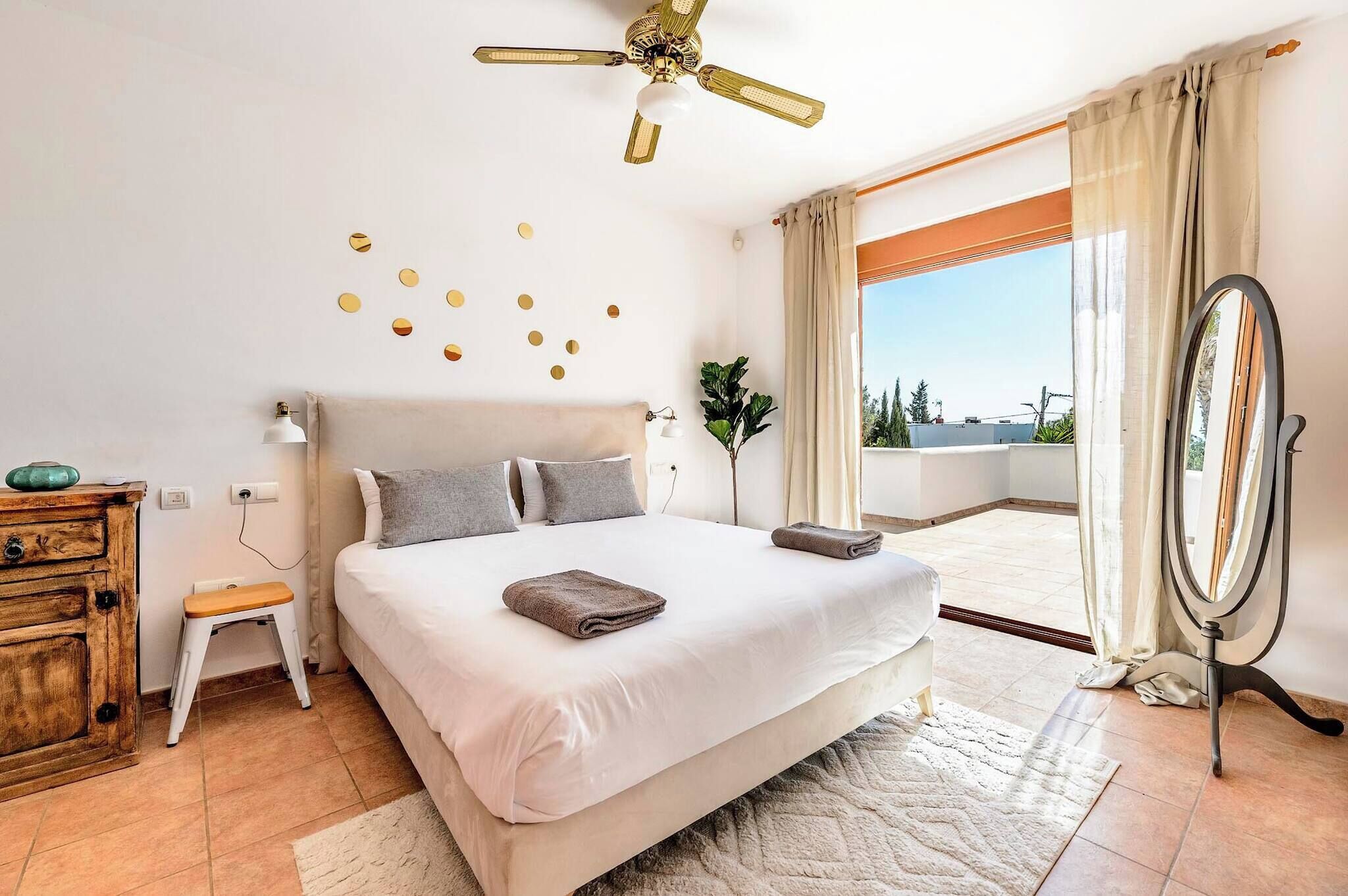Un dormitorio amplio con una cama grande, un ventilador de techo, un taburete pequeño, un aparador de madera, un espejo y vistas al exterior a través de un ventanal.