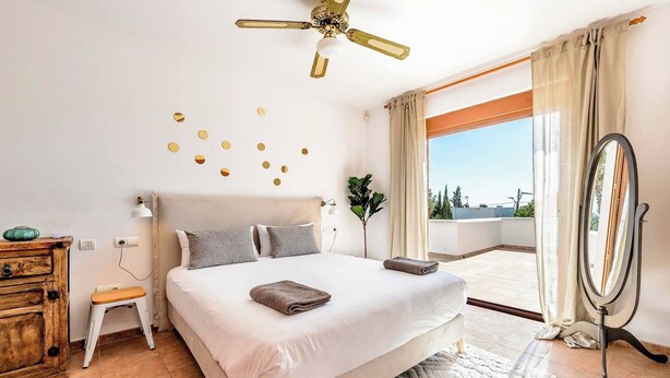 A spacious bedroom with a large bed, a ceiling fan, a small stool, a wooden dresser, a mirror, and a view of the outdoors through a large window. - Live the Ibiza Experience (Sant Josep de sa Talaia)
