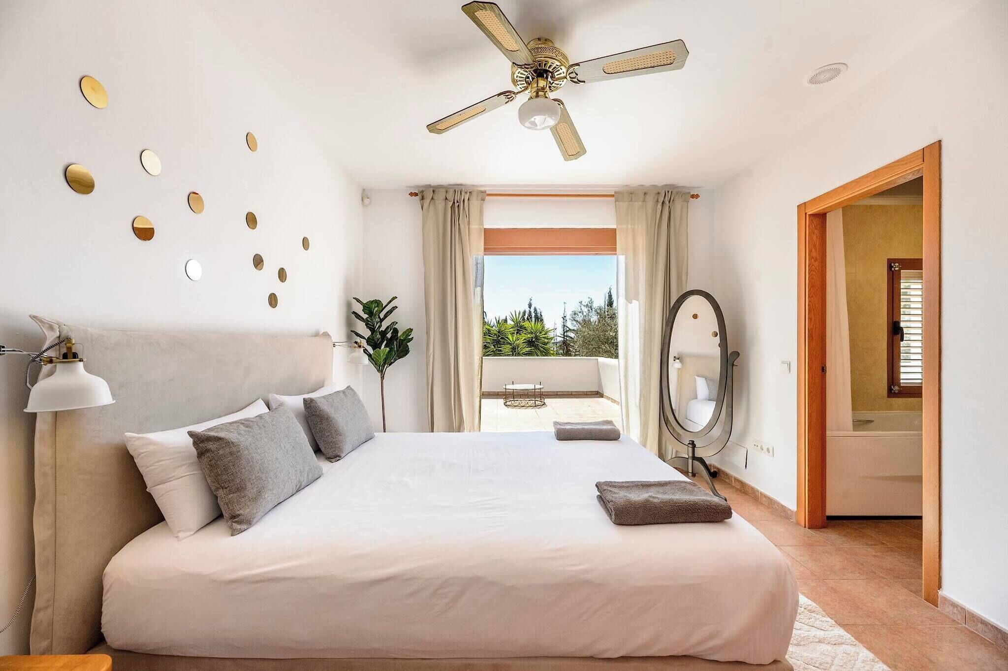 Un dormitorio con cama, ventilador de techo, ventana grande con cortinas, espejo y vistas al exterior.