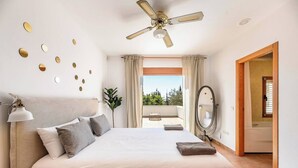 A bedroom with a bed, a ceiling fan, a large window with curtains, a mirror, and a view of the outdoors. - Live the Ibiza Experience (Sant Josep de sa Talaia)