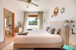 A bedroom with a large bed, a ceiling fan, a window with curtains, and a view of palm trees. - Live the Ibiza Experience (Sant Josep de sa Talaia)