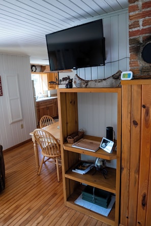 A wooden dining table with chairs, a television mounted on a brick wall, and a shelf with books and a clock.