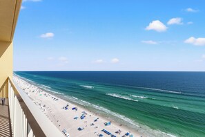 Blick auf einen Strand vom Balkon aus, mit blauem Himmel, Meereswellen und Strandstühlen.