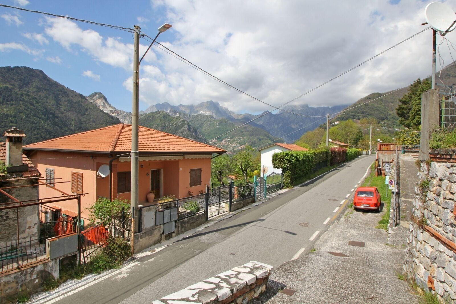 Una calle estrecha flanqueada por casas, un coche rojo estacionado a la derecha y montañas al fondo.