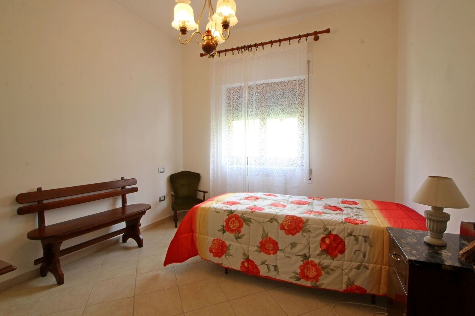 Une chambre à coucher avec un lit, un banc, une chaise, une lampe et une fenêtre avec des rideaux.