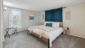 Ein Schlafzimmer mit Bett, kleinem Tisch, Stuhl und Fenster mit Jalousien und Vorhang.