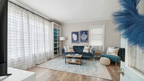 Ein Wohnzimmer mit einer Couch, einem Couchtisch und einem Fernseher. An der Wand hängen zwei gerahmte Bilder und eine blaue Zierpflanze.