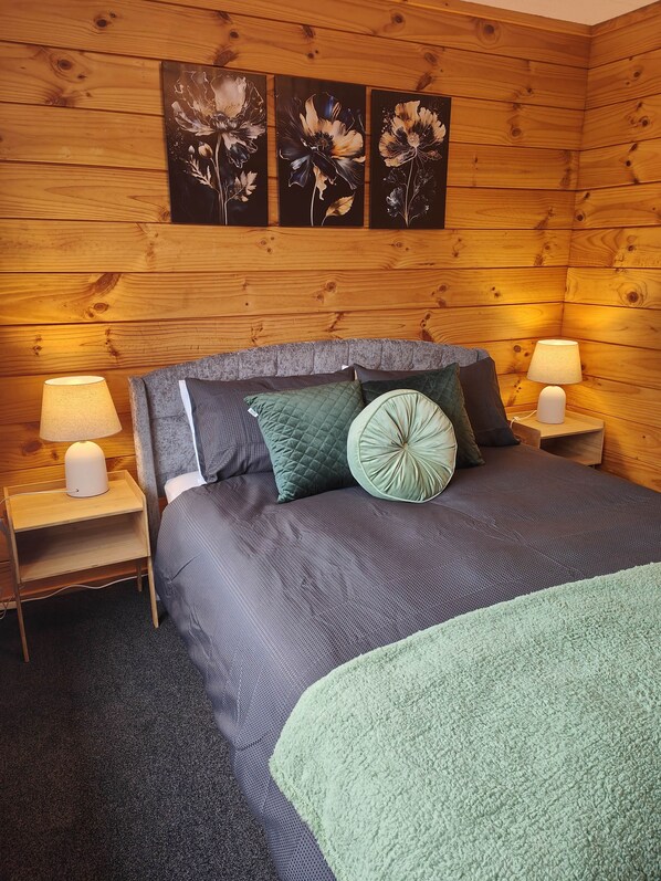 Ein Zimmer mit Holzwänden, einem Bett, zwei Nachttischlampen und drei floralen Bildern an der Wand.