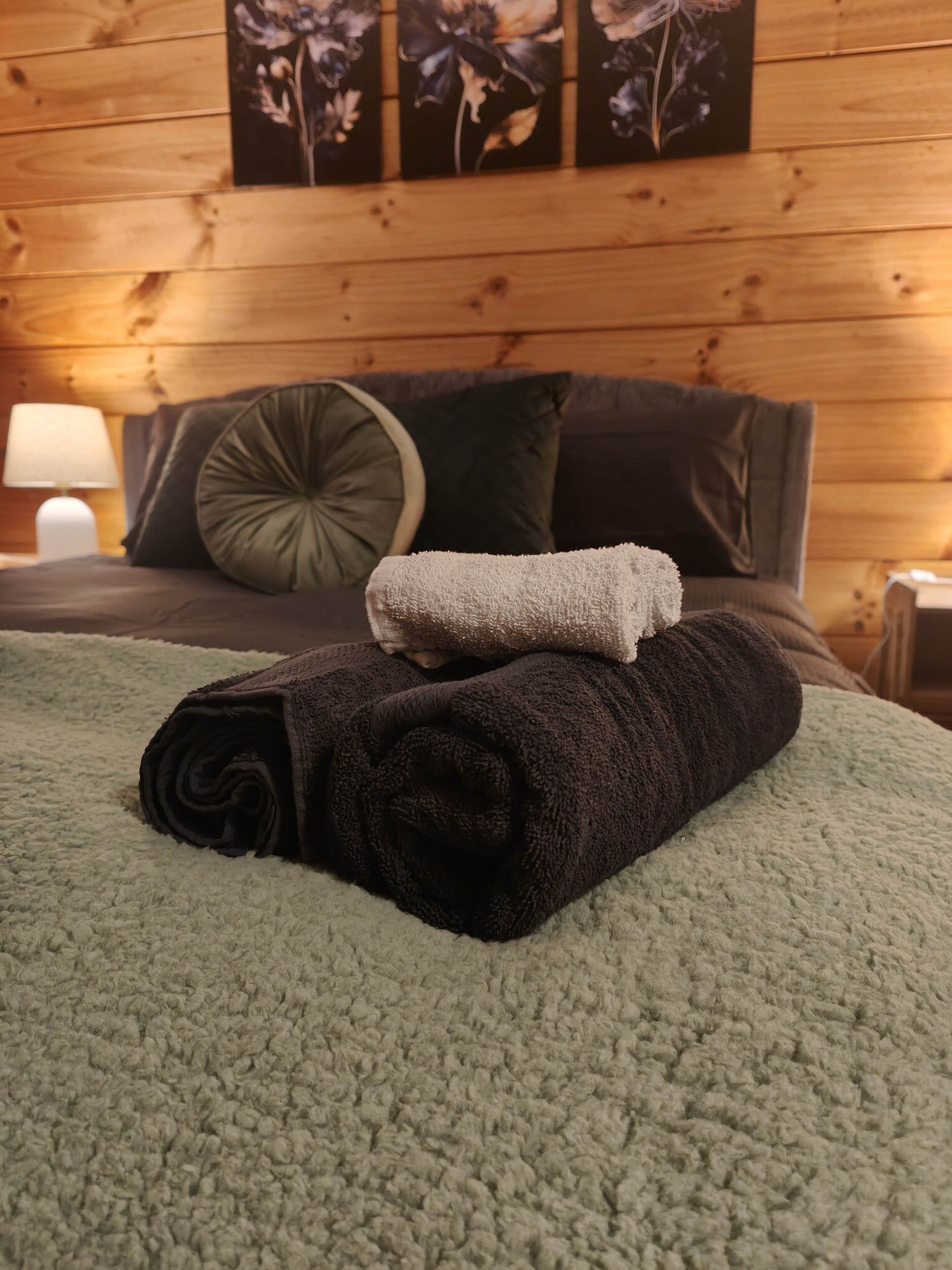 Un lit bien fait, avec une couverture verte et des serviettes enroulées, un oreiller marron foncé et un petit oreiller vert décoratif posé sur une tête de lit en bois.