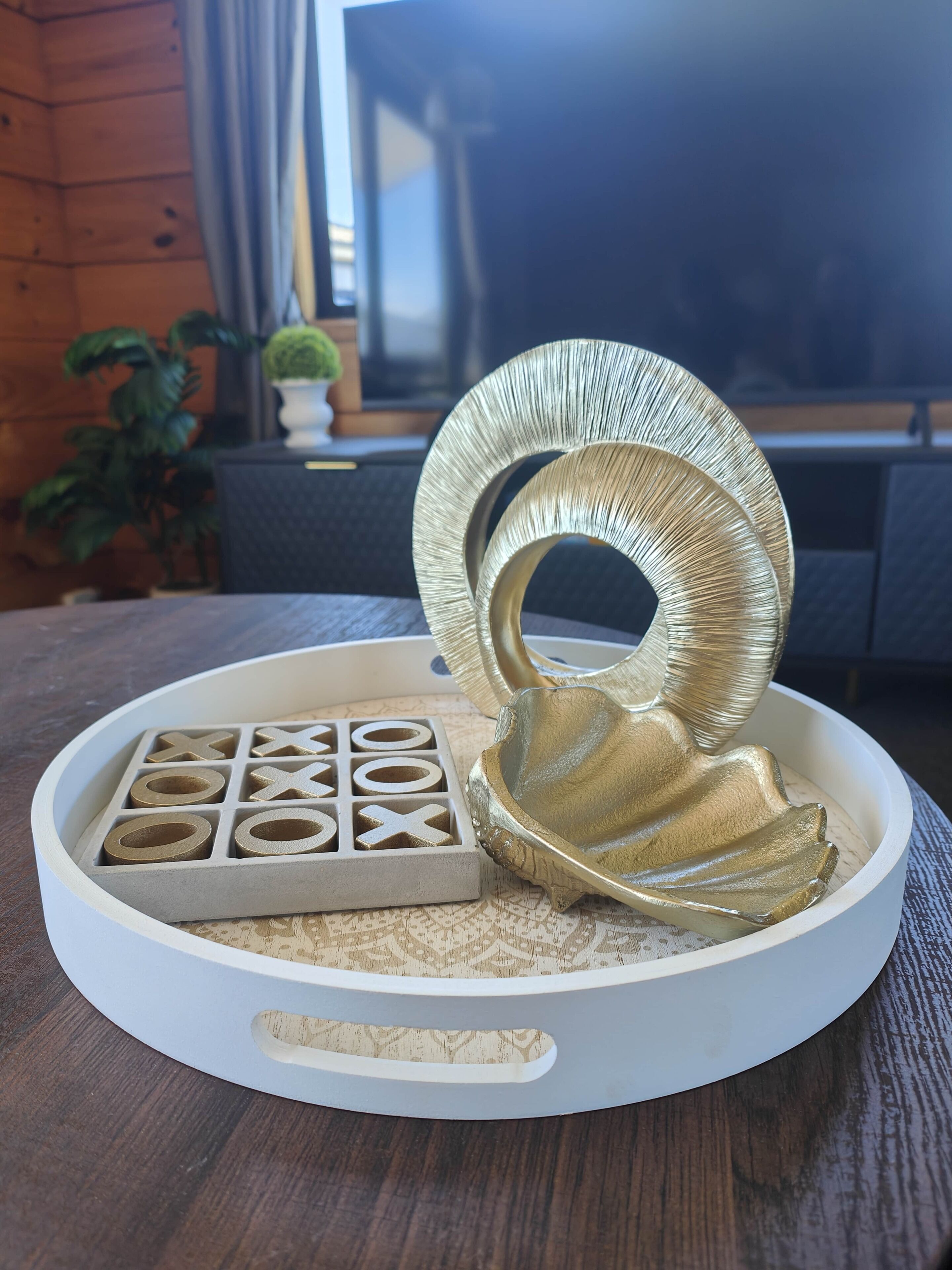 Uma mesa de madeira com um tabuleiro branco contendo um jogo de pedra, papel e tesoura e um objeto dourado decorativo.
