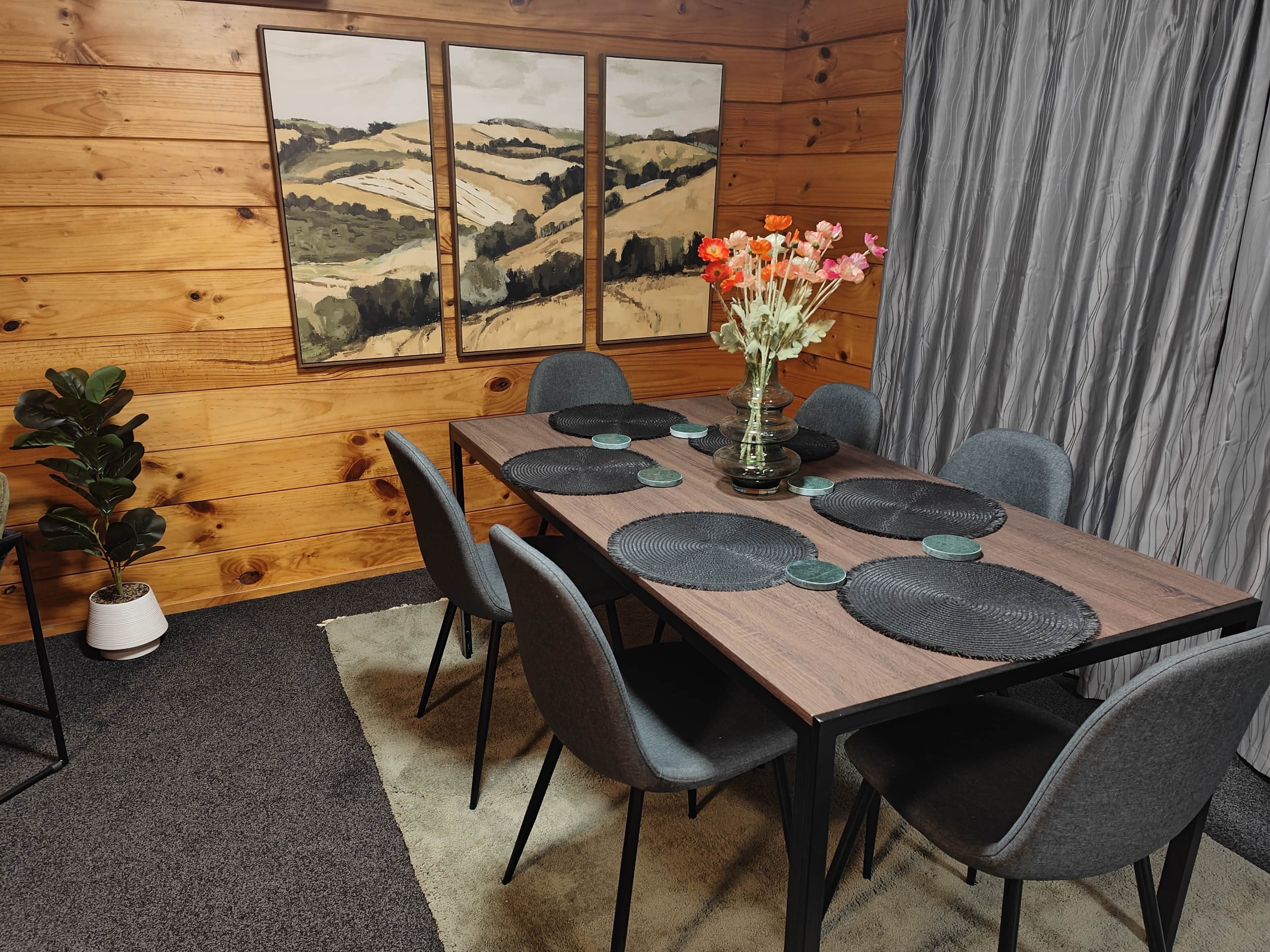 Une pièce aux murs lambrissés, avec une table à manger dressée pour six personnes, entourée de chaises grises. Un vase rempli de fleurs est posé sur la table, et trois tableaux représentant des paysages, encadrés, ornent le mur.
