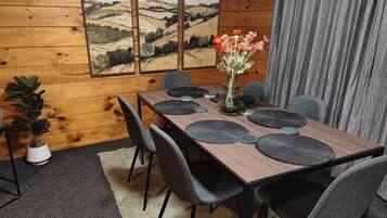 Une pièce aux murs lambrissés, avec une table à manger dressée pour six personnes, entourée de chaises grises. Un vase rempli de fleurs est posé sur la table, et trois tableaux représentant des paysages, encadrés, ornent le mur.