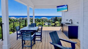 Een overdekt terras buiten met een tafel, stoelen, een plafondventilator en een televisie aan de muur.