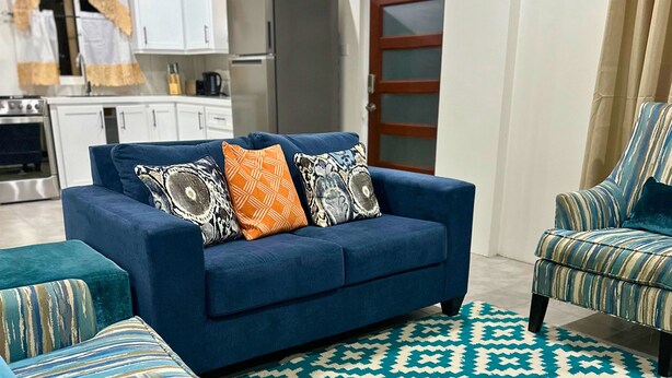 A living room with a blue sofa, matching armchair, and ottoman, a patterned rug, and a kitchen in the background. - Cozy 1-bedroom apartment in lovely Castries with AC (Castries)