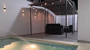 Une piscine avec un patio couvert, des meubles d’extérieur et des guirlandes lumineuses.