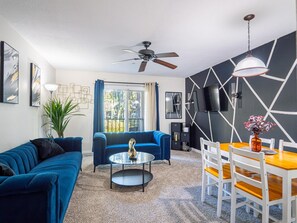 A modern living room with a blue sofa, a round coffee table, a dining table with chairs, a ceiling fan, and a TV mounted on the wall. - Cozy and Fun Family Bungalow: Pool+Gym+Near Disney World (Kissimmee)