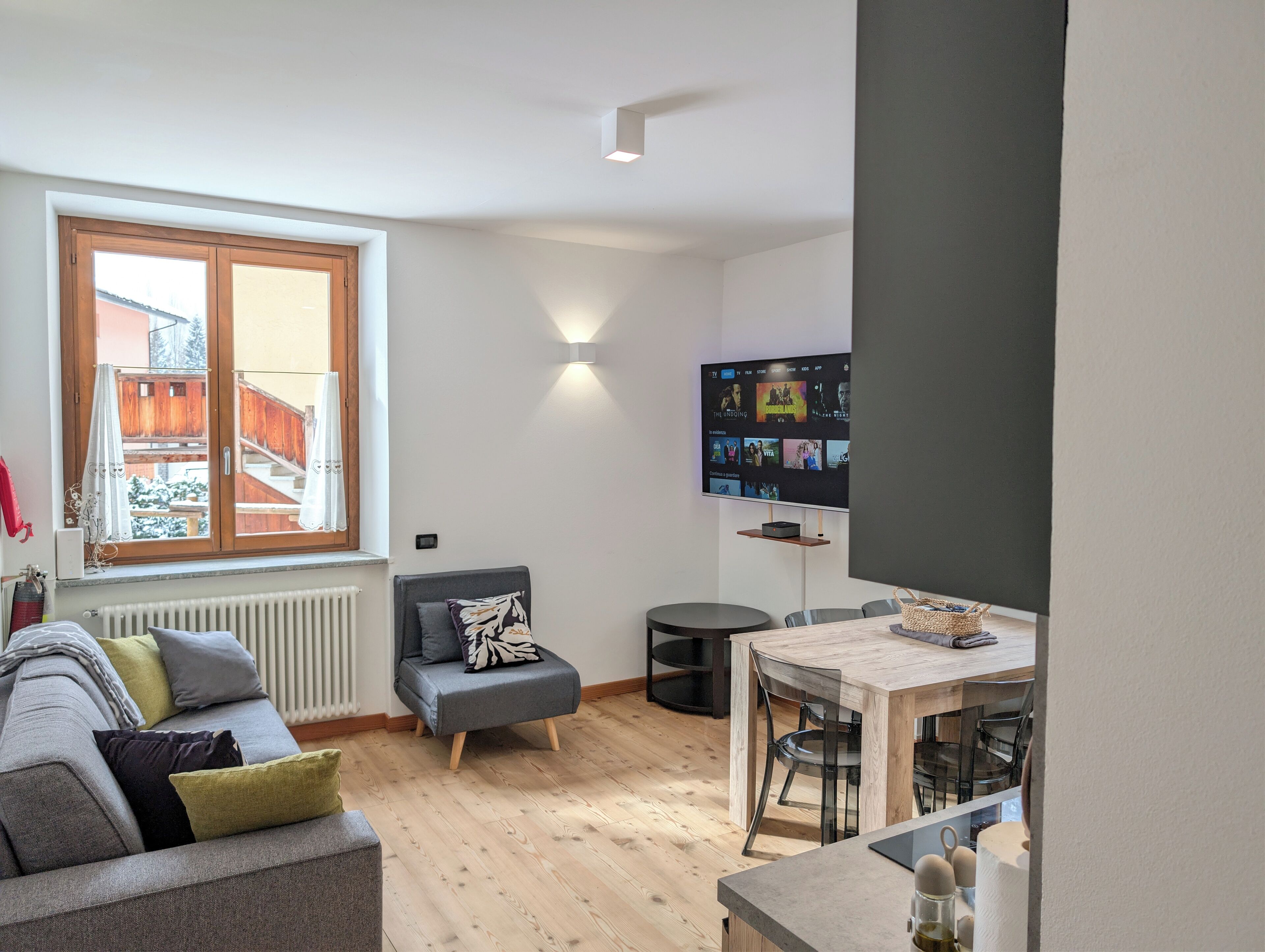 Een moderne woonkamer met een grijze bank, een klein grijze hocker, een houten eettafel met stoelen, een aan de muur bevestigde televisie en een raam met uitzicht op een balkon.