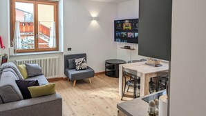 En moderne stue med en grå sofa, en lille grå skammel, et træspisebord med stole, et fjernsyn monteret på væggen og et vindue med udsigt til en balkon.