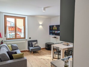 A modern living room with a grey sofa, a small grey ottoman, a wooden dining table with chairs, a TV mounted on the wall, and a window with a view of a balcony. - Ai piedi del Gigante apartment Cervinia-Valtournenche (Maen)