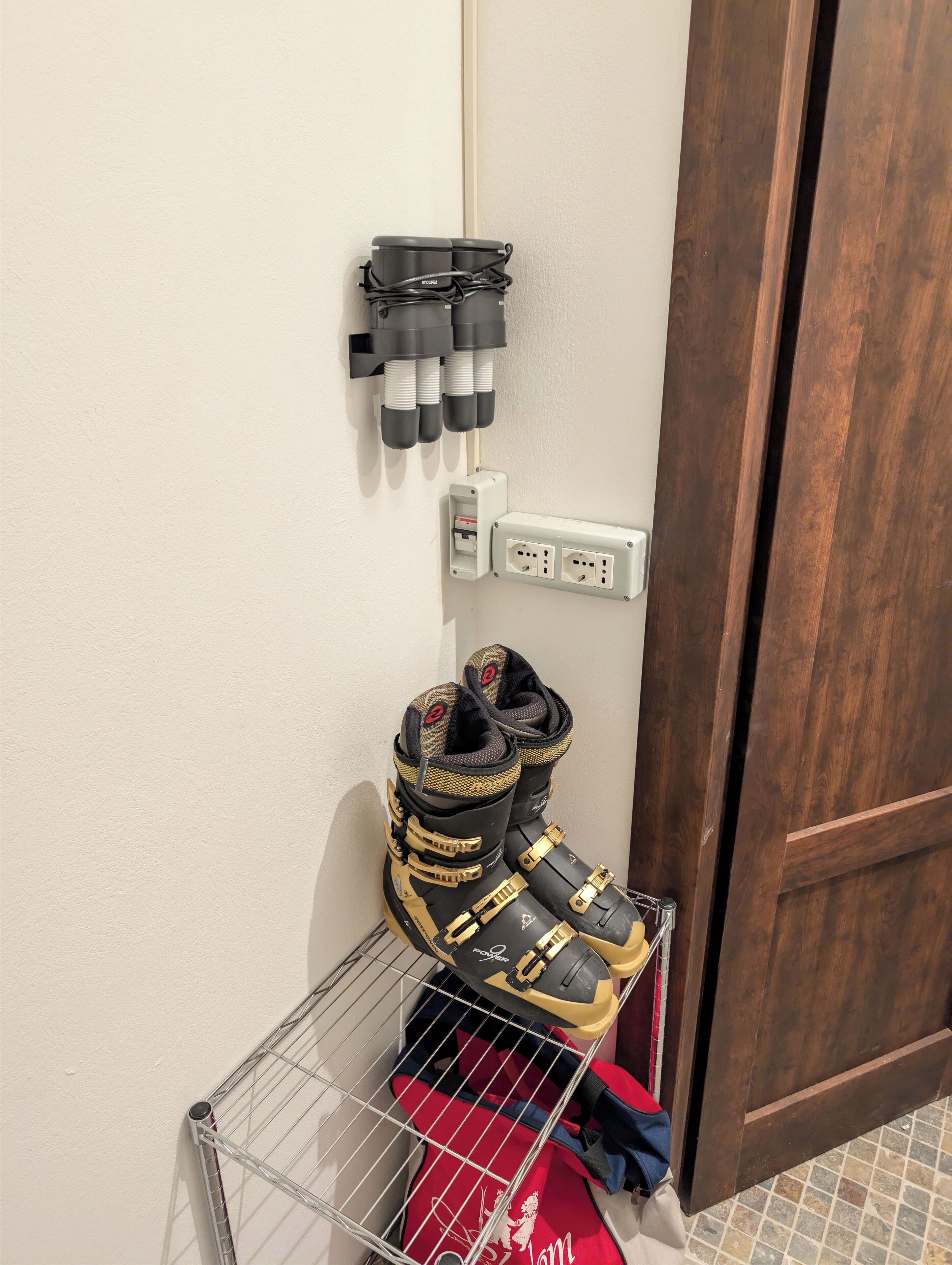Een hoek van een kamer met een deur, een wandcontactdoos en een metalen rek met een paar laarzen.