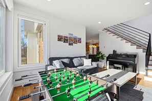 A modern living room with a foosball table, a sofa, a piano, and a staircase. - Sleeps 10 • New Modern Home by Swamp Rabbit Trail (Greenville)