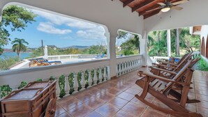 Un espace extérieur abrité, avec vue sur la mer, des fauteuils à bascule en bois et une balustrade blanche.