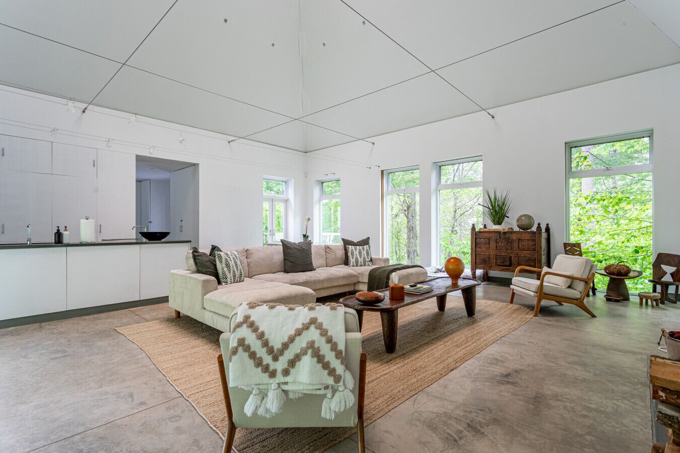 A spacious living room with a large sofa, a coffee table, and a dining area. There are large windows providing natural light and a view of greenery outside.