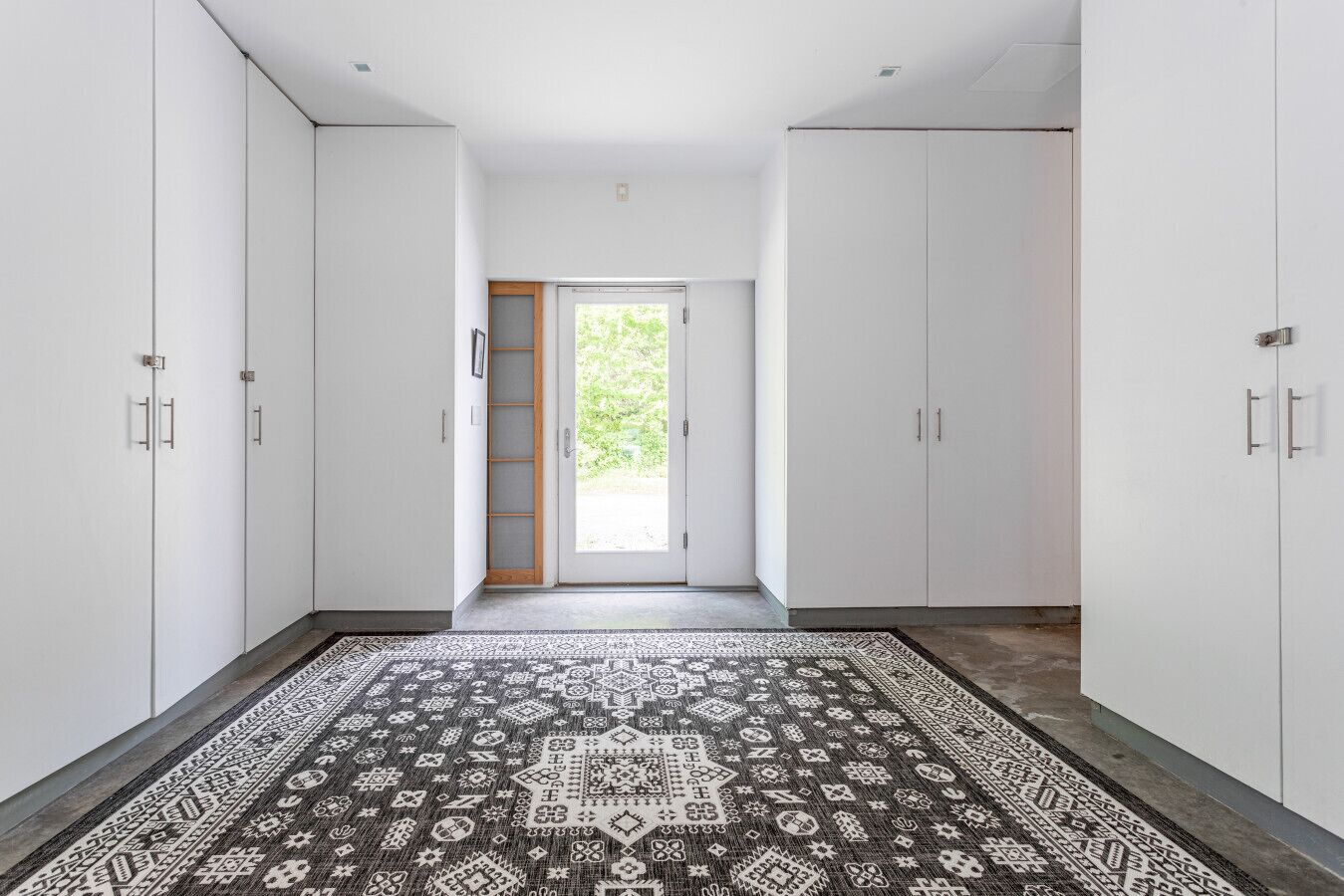 Un couloir avec des armoires blanches, un tapis à motifs et une porte donnant sur l’extérieur.