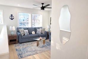 Sala de estar con sofá gris, cojines estampados, una mesa de centro de vidrio y una alfombra azul. Hay dos mesitas de noche con lámparas, un ventilador de techo y un ventanal con persianas.