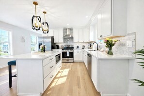 Une cuisine moderne avec des meubles blancs, un îlot central, des appareils en acier inoxydable et des luminaires suspendus.