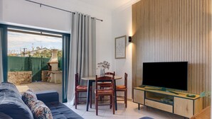 Un salon comprenant un canapé, un téléviseur, un mur recouvert de panneaux en bois, une table à manger avec des chaises, et une vue à travers une porte vitrée.