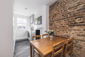 A dining area with a wooden table and chairs, a brick wall, and a kitchen in the background. - The Victorian Maisonette – Timeless Elegance in the Heart of London (London)