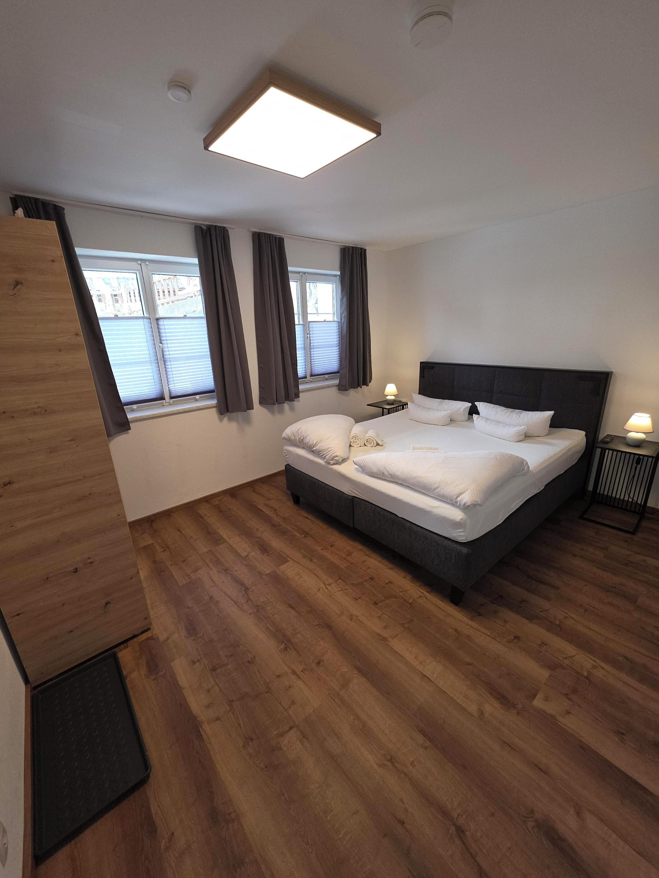 A modern bedroom with a large bed, a bedside table, a nightstand, a ceiling light, and a window with curtains.